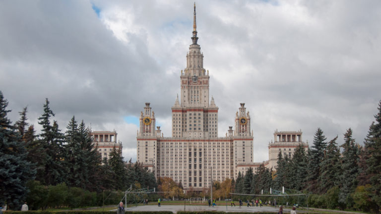 Moscow State University seen beyond lawns and evergreen trees, with its central spire and flanking clock towers.
