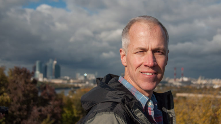 Man in a dark jacket standing outdoors with the blurred Moscow City skyline and autumn landscape behind him.