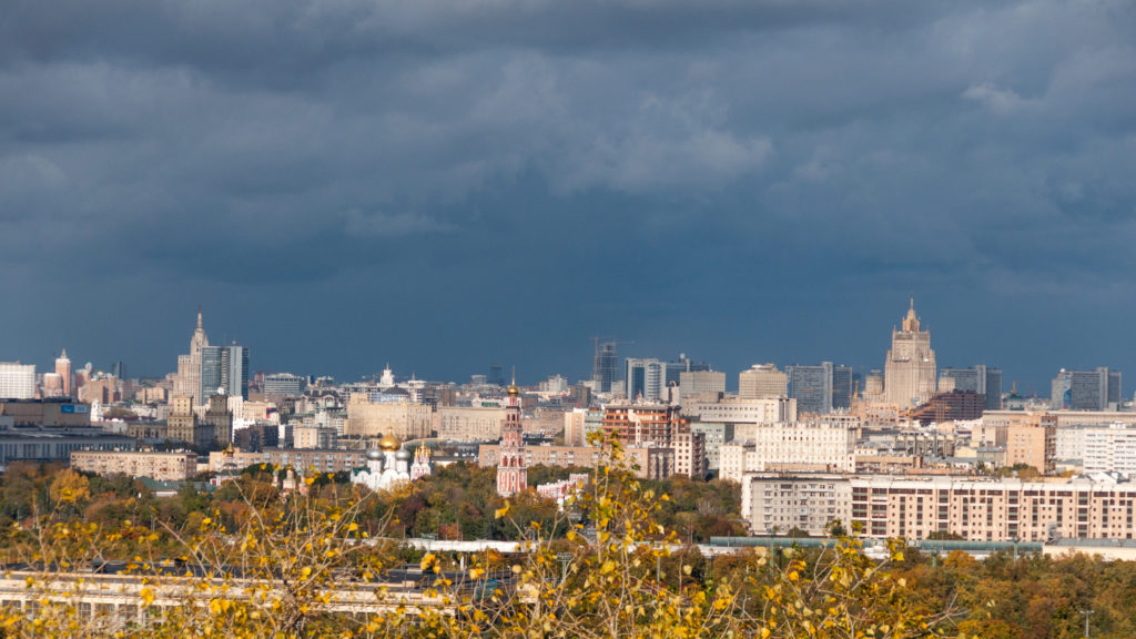 Wide Moscow skyline with monumental pale buildings beneath dark storm clouds and autumn trees in the foreground.