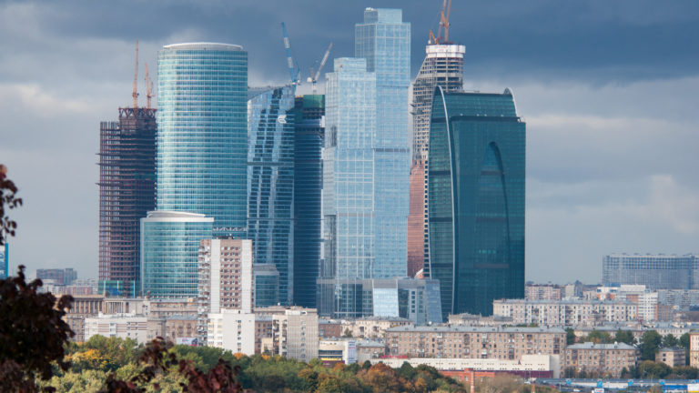 Cluster of glass Moscow City skyscrapers, several under construction with cranes, rising above lower urban buildings.