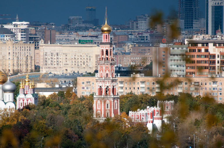 Red-and-white Novodevichy Convent bell tower with gilded dome rising above autumn trees and apartment buildings.