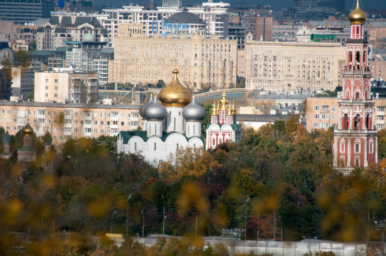 Novodevichy Convent domes and red-and-white bell tower rising above autumn trees with city buildings behind.