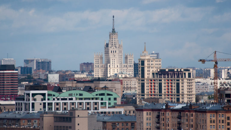 Spired Stalinist high-rise rising beyond mixed Moscow buildings and construction cranes in a telephoto skyline view.