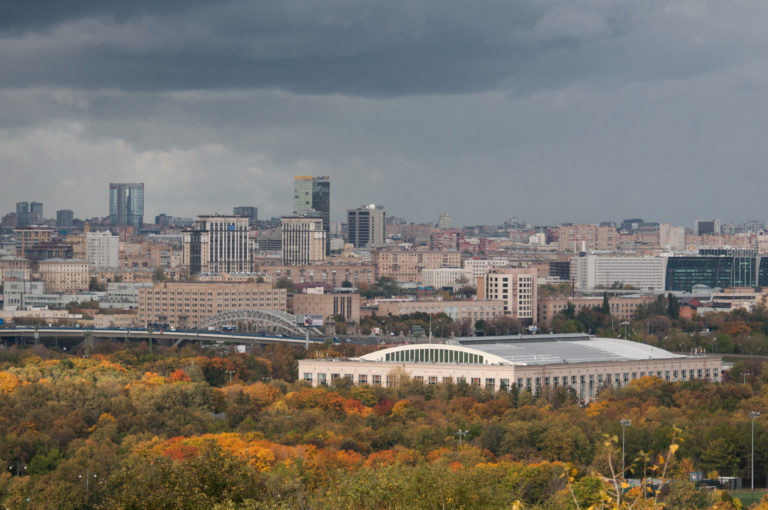 Wide Moscow skyline beneath dark clouds, with autumn woodland and a large pale-roofed building in the middle distance.
