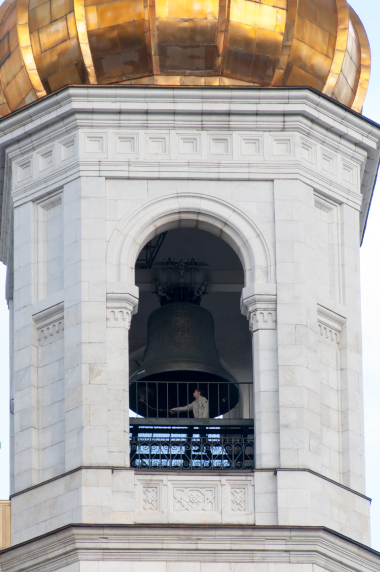 Large dark bell hanging inside an arched white tower opening beneath a gilded roof at the cathedral.
