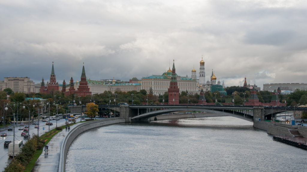 Moscow Kremlin walls, red towers, green-roofed buildings, and gold domes seen across the Moscow River and bridges.