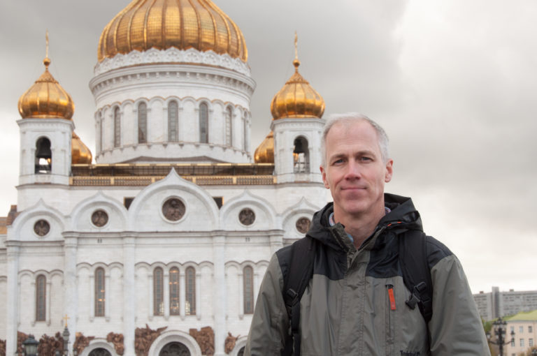 Man with backpack standing before the white façade and gilded domes of the Cathedral of Christ the Saviour.