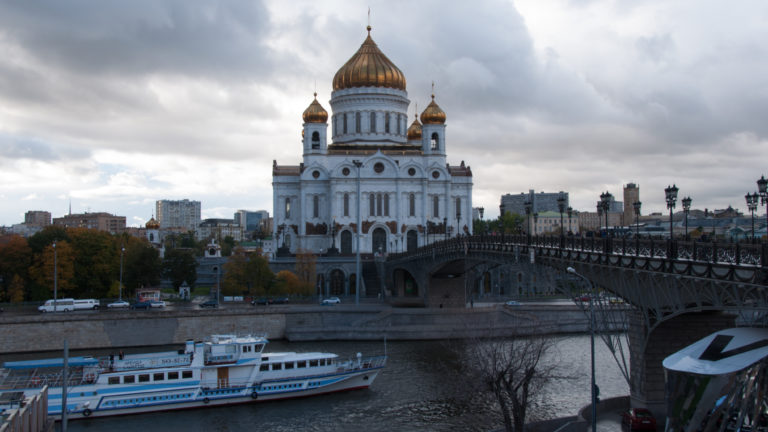 White-and-gold Cathedral of Christ the Saviour beside a curving bridge, with a passenger boat on the Moscow River.