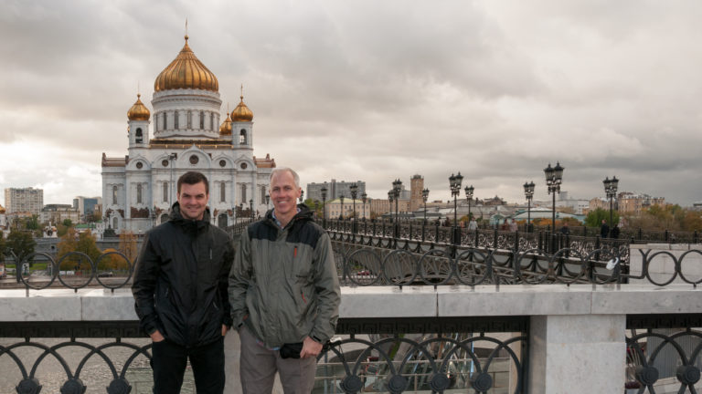 Two men standing at a bridge railing with the Cathedral of Christ the Saviour centered behind them.