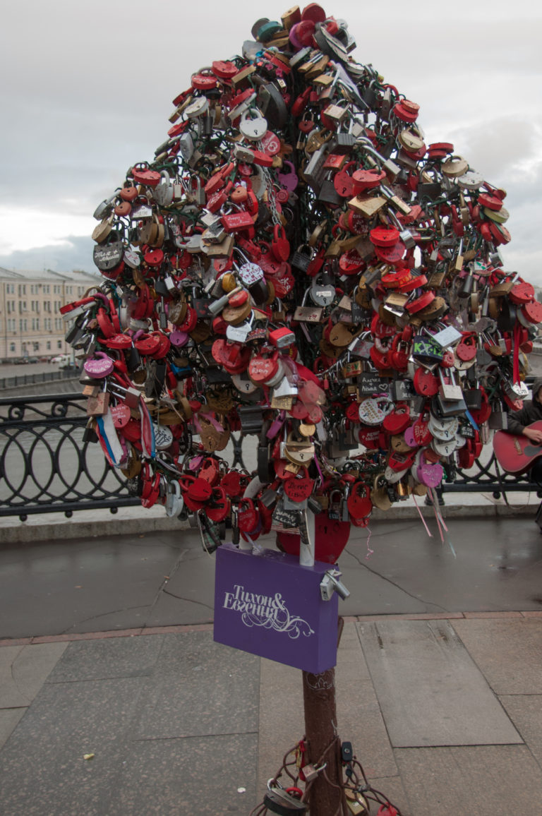 Tree-shaped metal installation densely covered with red and multicolored love locks beside a Moscow river railing.