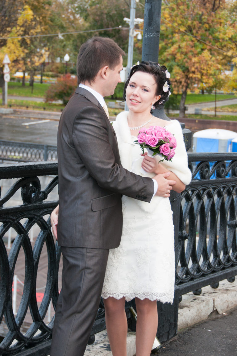 Bride with pink bouquet and groom standing beside an ornamental iron railing on a Moscow river embankment.