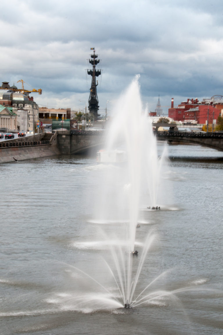 River fountain spraying in front of the distant Peter the Great monument and waterfront buildings under gray clouds.
