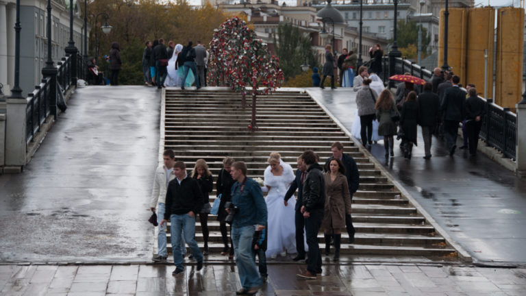 Bride and groom with companions near broad wet steps as pedestrians pass on a rainy Moscow walkway.