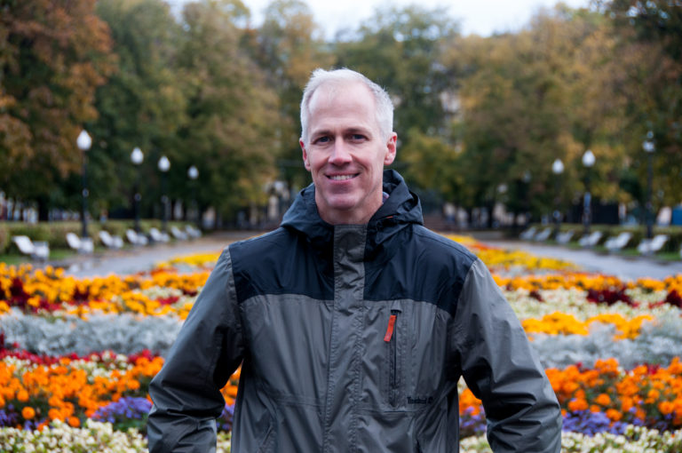 Smiling male visitor in a dark jacket standing before colorful autumn flower beds in a tree-lined park.
