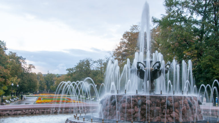 Tiered park fountain with tall central jet, arcing side jets, flower beds, and tree-lined paths.