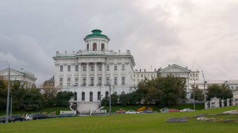White classical multi-story building with tall windows and a green-capped rooftop rotunda beyond a broad lawn.