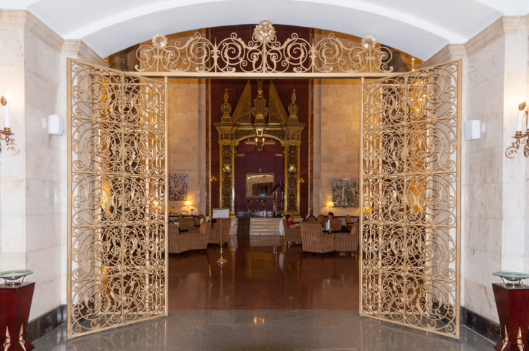 Open gilded scrollwork gates framing a polished formal hall with symmetrical furnishings beyond.