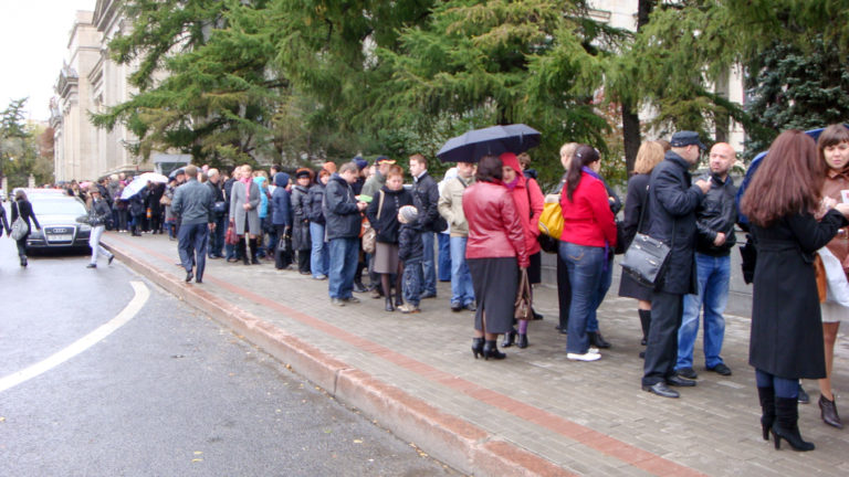 Long line of people in coats and umbrellas waiting along a wet city sidewalk beside the street.