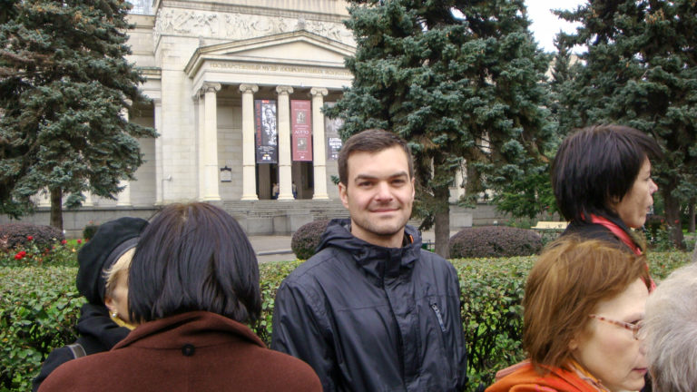 Smiling male visitor among a tour group, with a columned stone building and evergreen trees behind him.