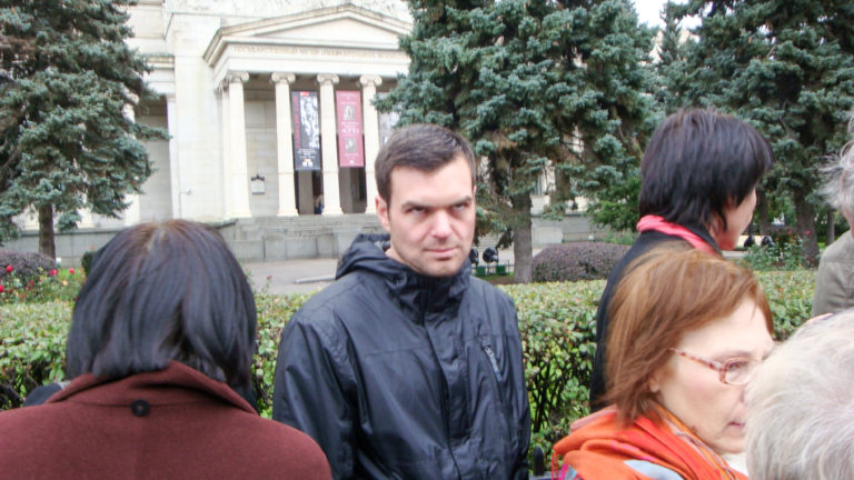 Several visitors in jackets and scarves stand before a landscaped plaza and large columned stone building.