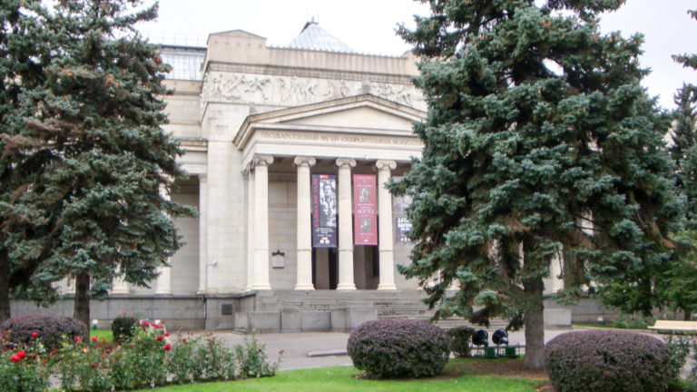Neoclassical stone museum facade with tall columned portico, exhibition banners, evergreens, shrubs, and flower beds.