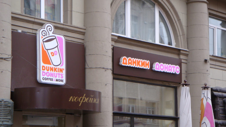 Dunkin' Donuts storefront on a stone building with English logo and illuminated Cyrillic brand signage.