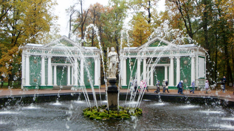 Ring fountain around a central statue before an ornate green-and-white pavilion among autumn trees.