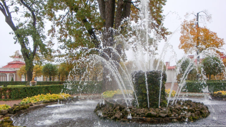 Rockwork fountain spraying multiple jets amid flowerbeds and autumn trees in Peterhof’s park.