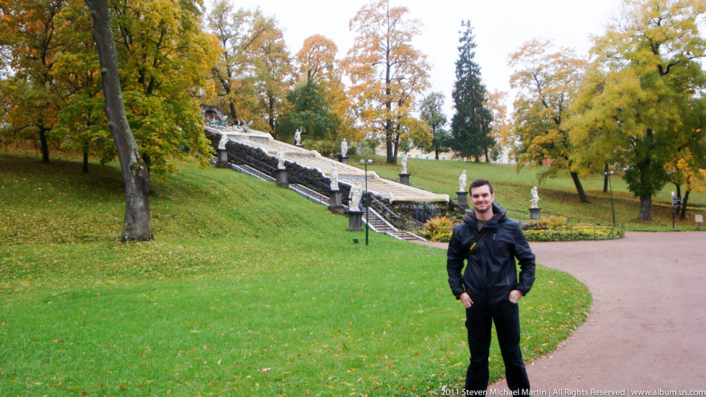 Man standing on a park path with autumn trees and a stepped water feature behind him.