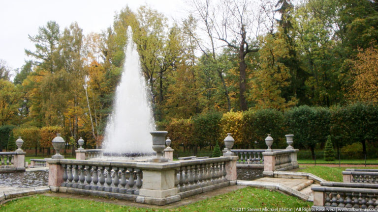 Tall fountain jet rising inside a stone-balustraded basin surrounded by autumn trees.