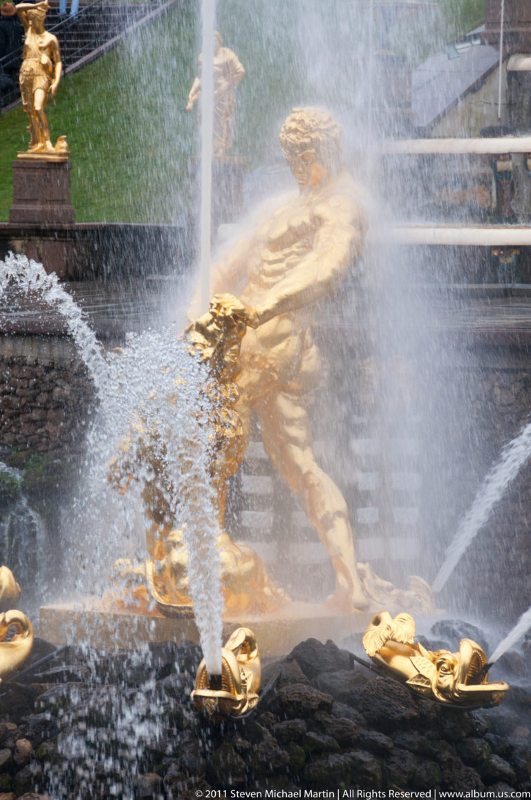 Reclining gilded fountain figure behind crossing water jets, mist, and small gilded spouts.