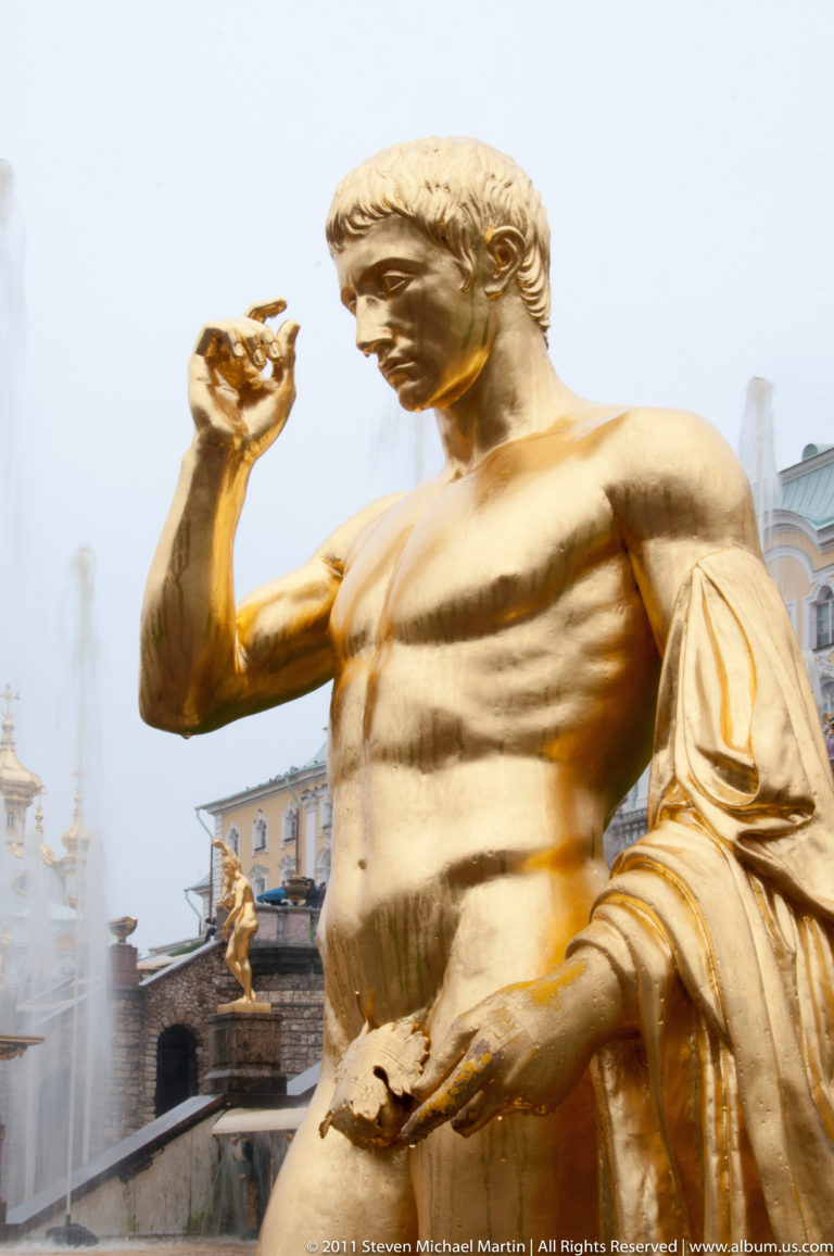 Close view of a gilded male fountain sculpture with water spray and palace architecture behind.