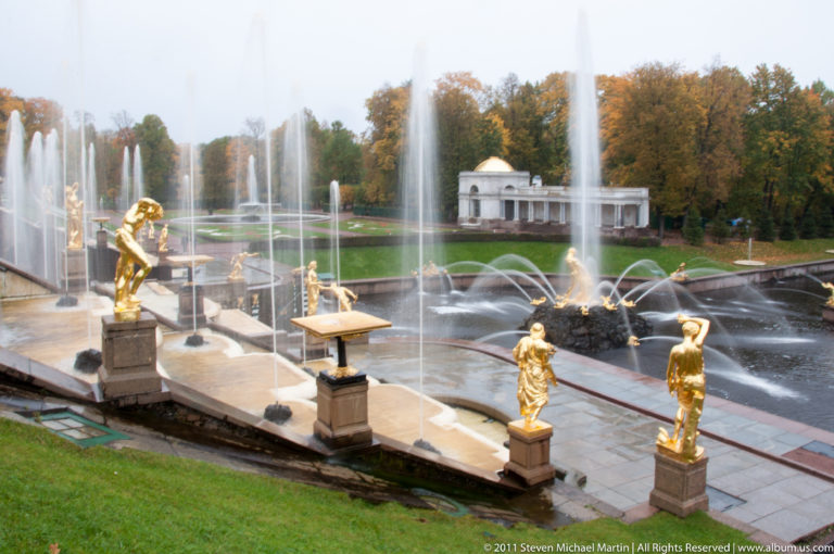 Gilded statues beside a fountain basin with tall jets, patterned lawns, pavilion, and autumn trees beyond.