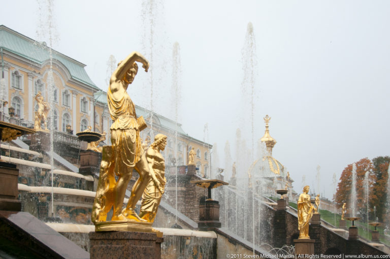 Gilded female fountain statue with raised arm beside Grand Cascade steps, jets, and palace architecture.