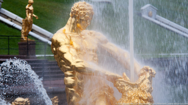 Close gilded muscular male fountain figure gripping an ornate form while water sprays across his torso.
