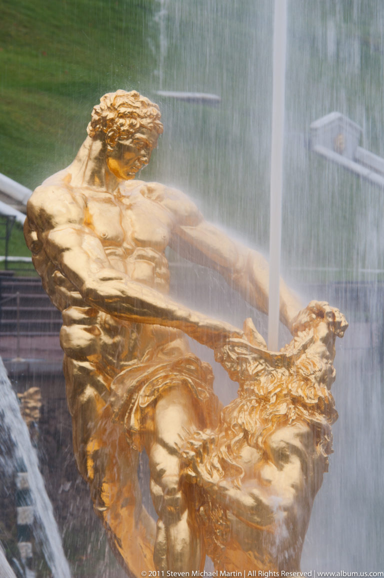Reclining gilded male fountain figure twisting beside an ornate element amid vertical and diagonal water jets.