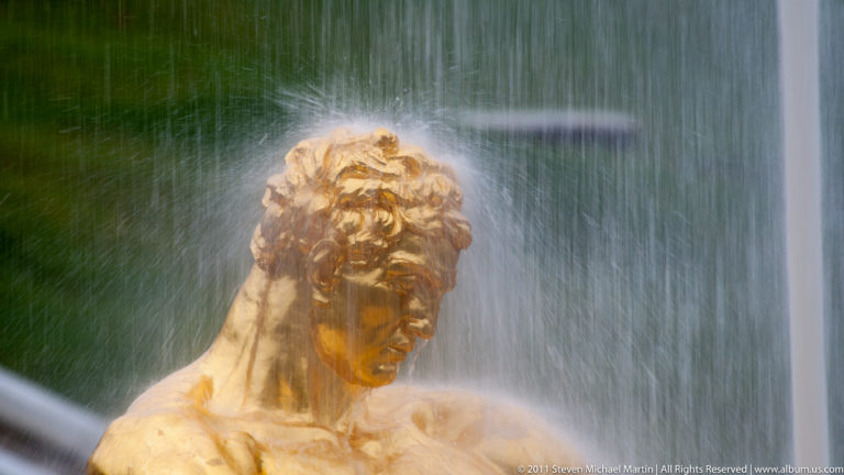 Head and shoulders of a gilded male fountain figure seen through a dense curtain of water.