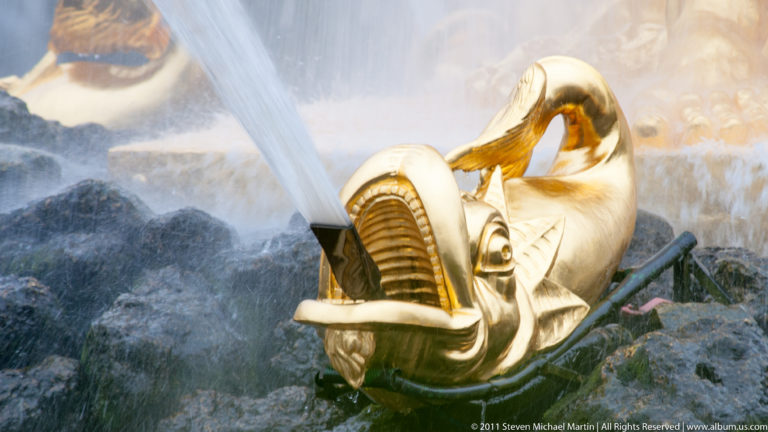 Gilded fish-like fountain spout with open mouth directing a strong jet over dark rockwork.