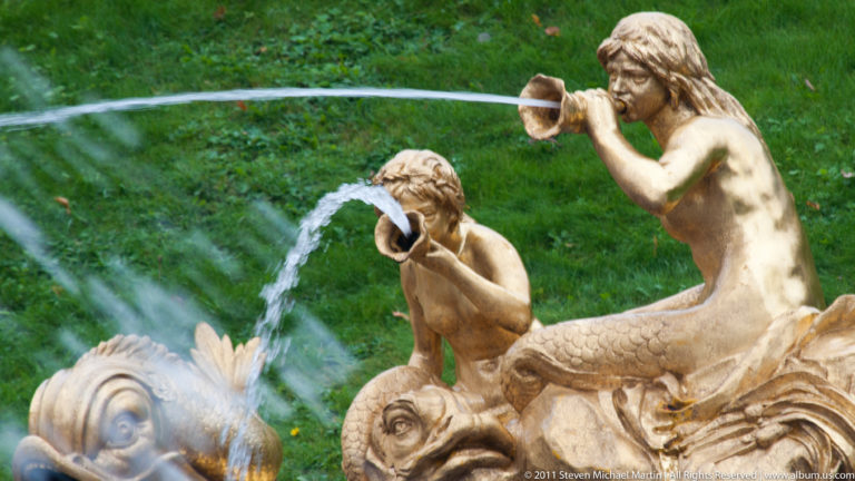 Two gilded fish-tailed figures blowing shell-like horns while fountain streams arc around them.