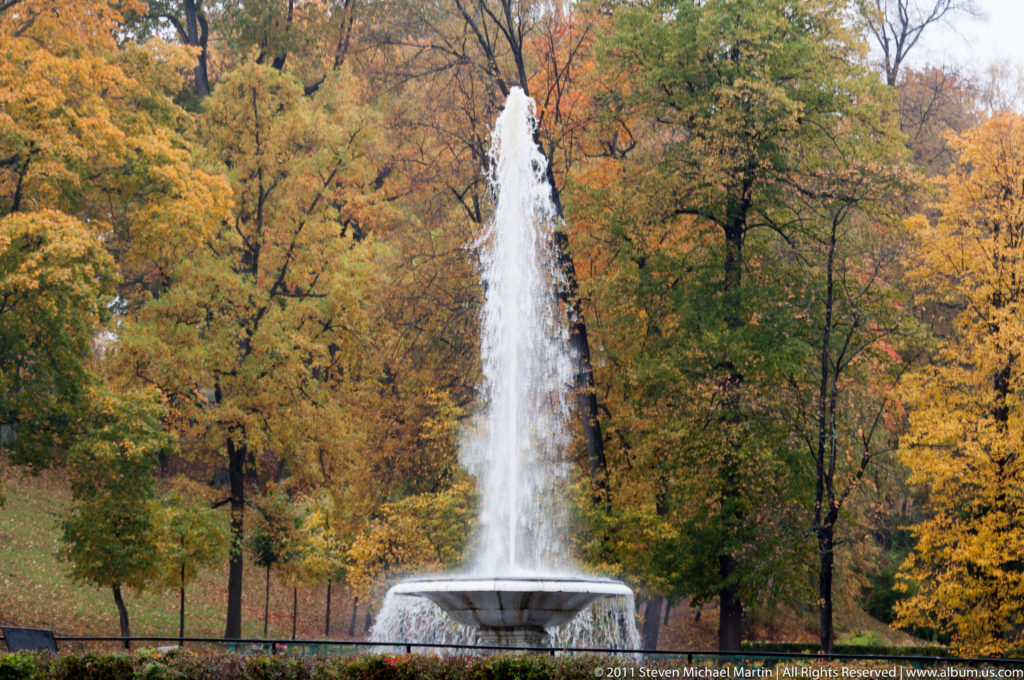 Tall fountain jet rising from a circular basin among yellow and orange autumn trees.