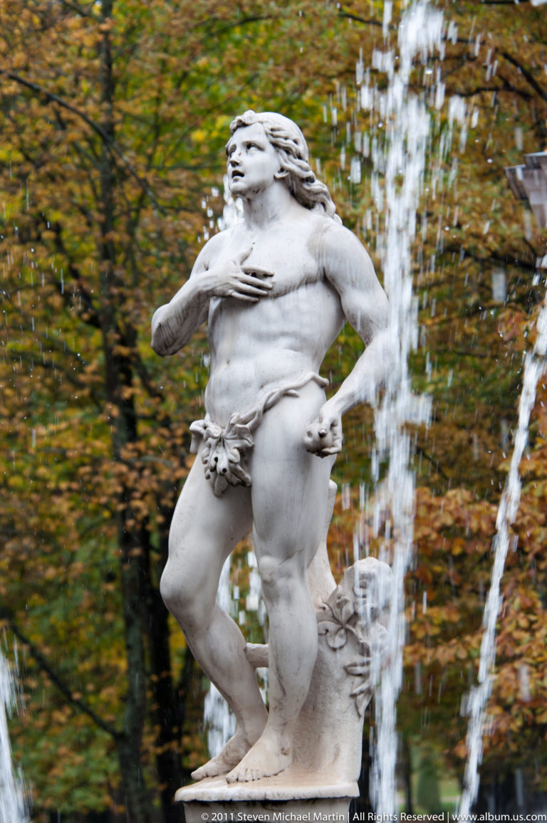 Pale classical male statue posed beside vertical fountain jets with autumn trees behind.