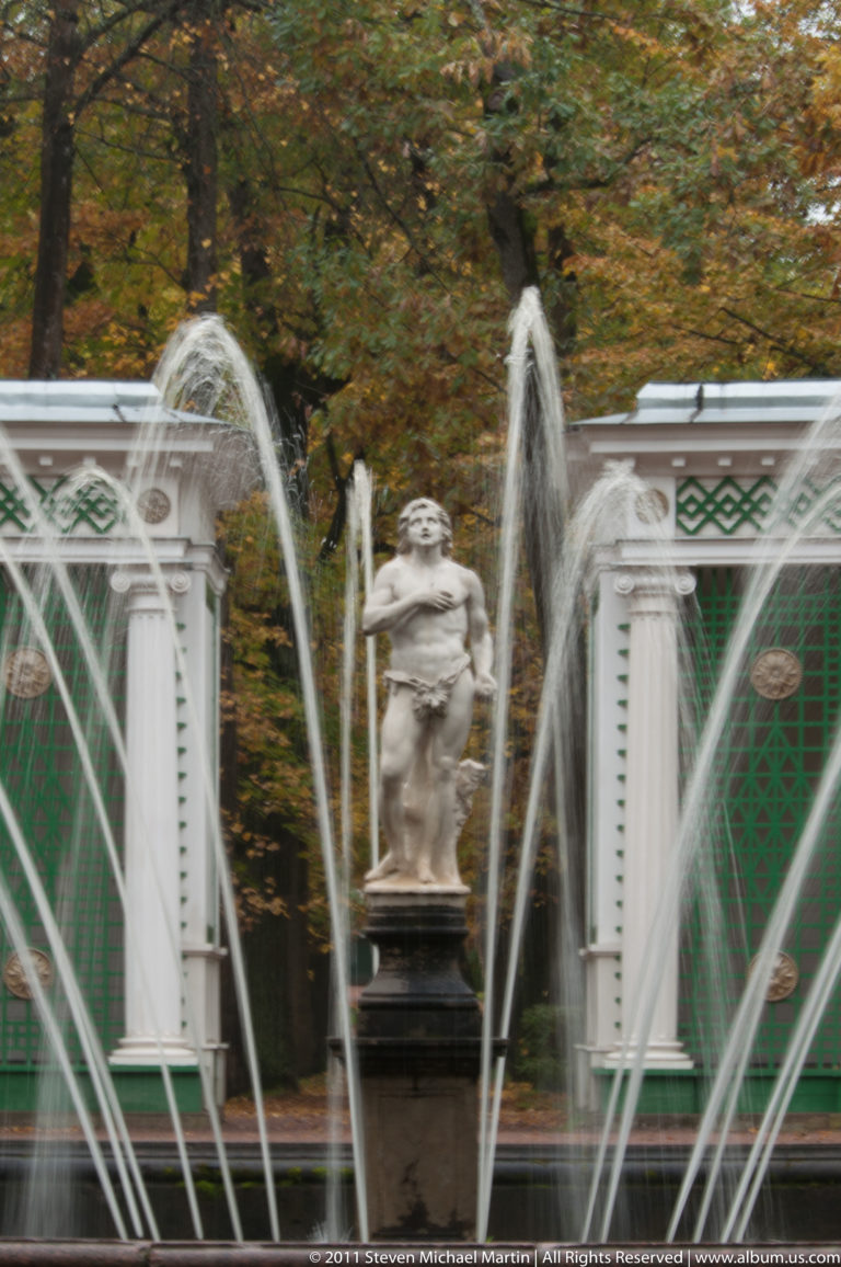 Classical statue on a pedestal framed by crossing fountain arcs before a green-and-white garden structure.