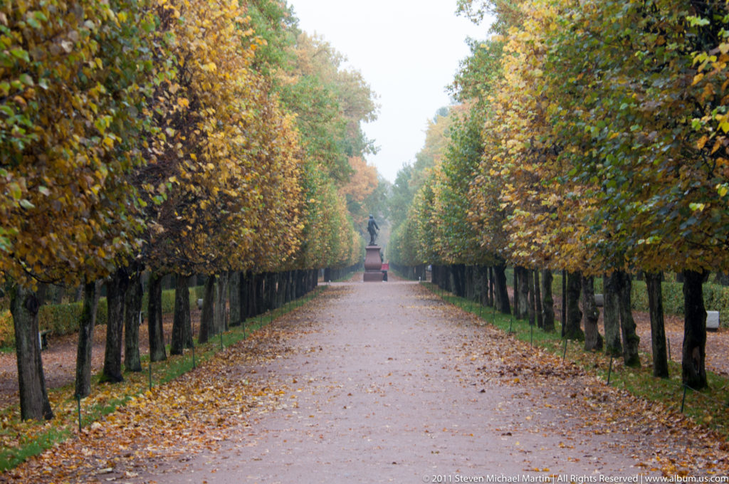 Straight gravel avenue lined by clipped trees and hedges, with autumn leaves and a distant garden feature.
