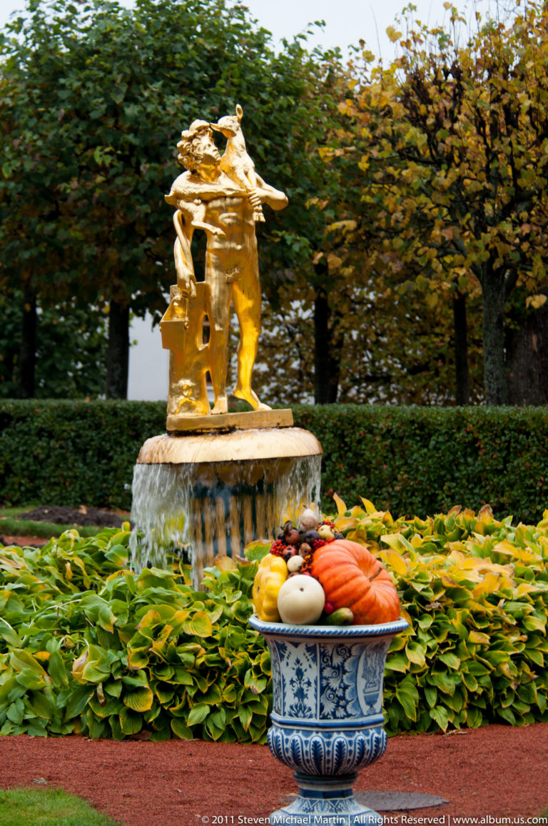Gilded sculptural fountain behind a blue-and-white pot filled with pumpkins and gourds in a formal garden.