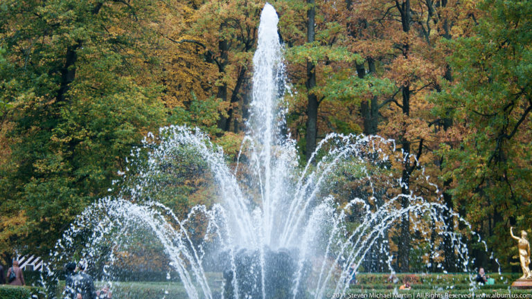 Tall central fountain jet with arcing side jets before autumn trees in Peterhof Lower Park.