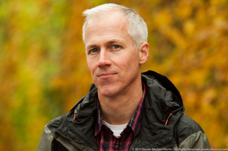 Close portrait of a gray-haired visitor in a dark jacket against blurred golden autumn foliage.