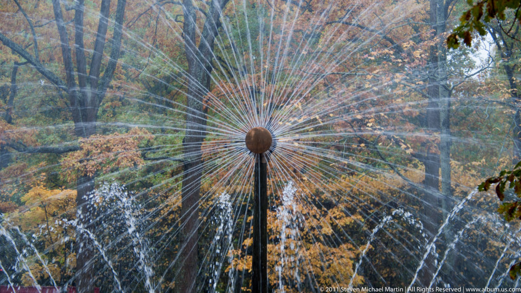 Circular radial fountain spraying many fine jets outward like spokes against autumn trees.