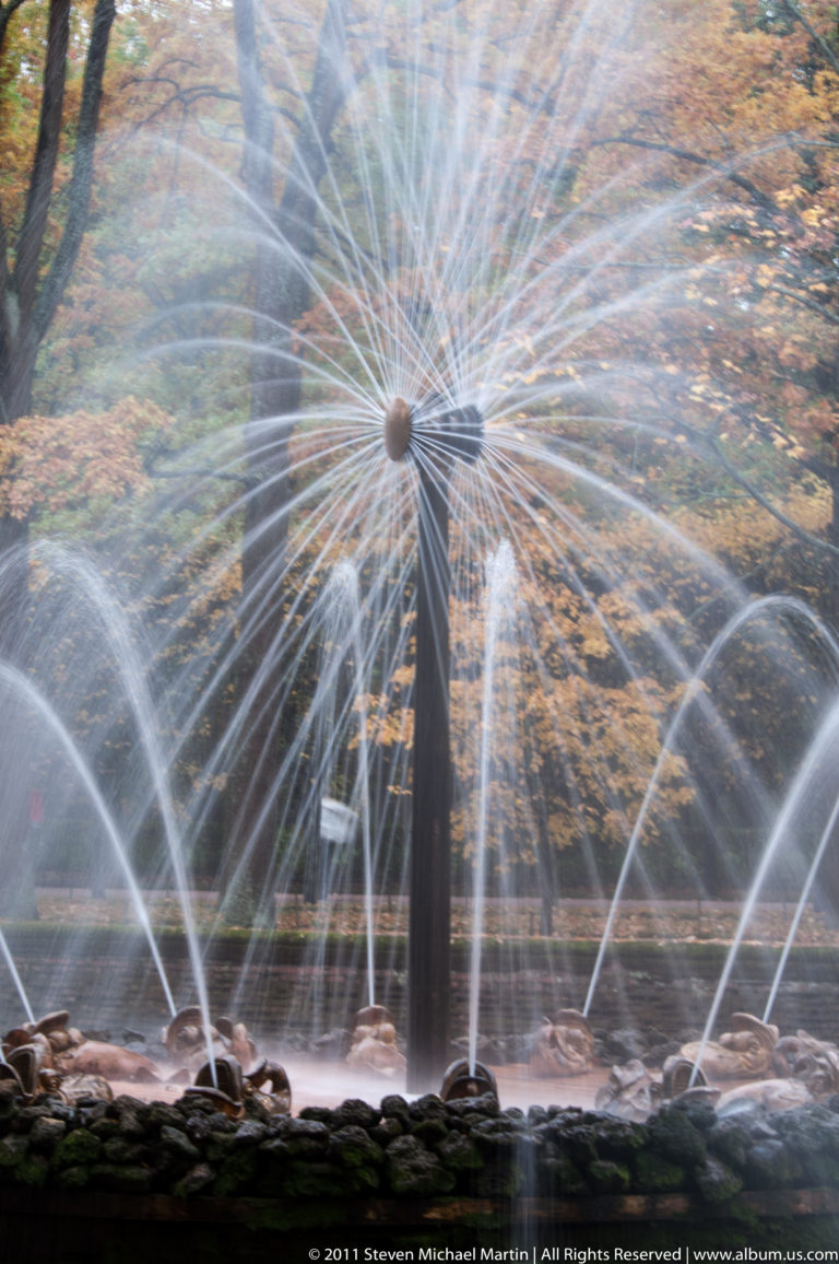 Wide radial fountain with spoke-like jets, larger arcing sprays, and dark rocks along the basin.