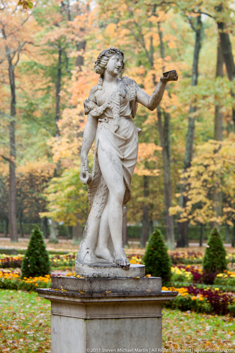 Classical female statue on a pedestal with one arm extended amid flower beds and autumn trees.