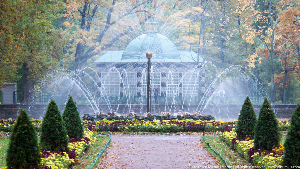 Radial fountain centered before a light garden pavilion, framed by conical evergreens and flower beds.